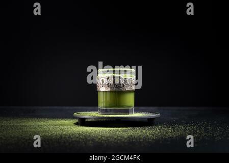 Gesunder grüner Matcha-Tee aus Kräutern, serviert in einer Glastasse mit Metalldekor auf einer Untertasse, die mit Pulver auf dem schwarzen Tisch bestreut ist Stockfoto
