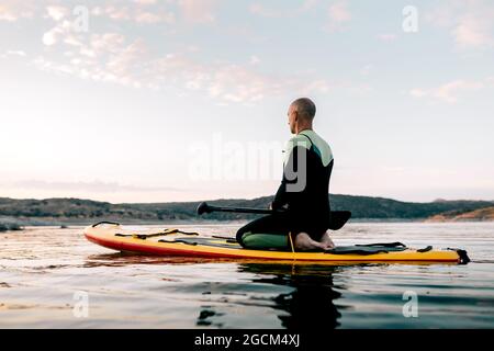 Seitenansicht des ruhigen männlichen Surfers, der in Thunderbolt-Pose auf dem Paddleboard sitzt und am Abend beim Yoga im Meer mediiert Stockfoto