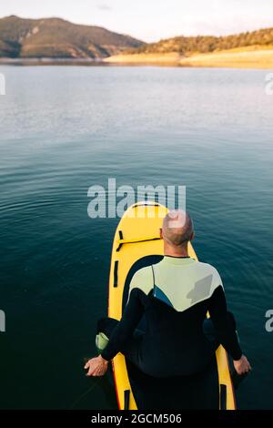 Von oben Rückansicht eines erwachsenen Mannes im Neoprenanzug, der auf dem Paddelbrett auf der ruhigen Wasseroberfläche des Sees kniet Stockfoto