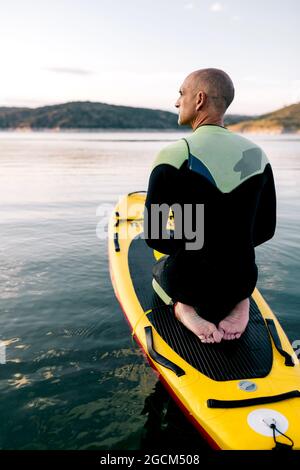Rückansicht des erwachsenen Mannes im Neoprenanzug, der auf dem Paddelbrett auf der ruhigen Wasseroberfläche des Sees kniet Stockfoto