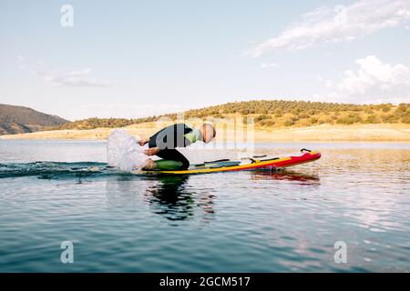 Ganzkörper-Seitenansicht des erwachsenen Mannes im Neoprenanzug, der auf dem Paddle-Board kniet und mit den Armen auf der ruhigen Wasseroberfläche des Sees paddelt Stockfoto