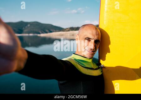 Positiv aktiver Mann mittleren Alters im Neoprenanzug, der in der Nähe des Paddle Boards steht und die Kamera anschaut, während er im Sommer da ein Selbstporträt gegen einen ruhigen See aufnahm Stockfoto