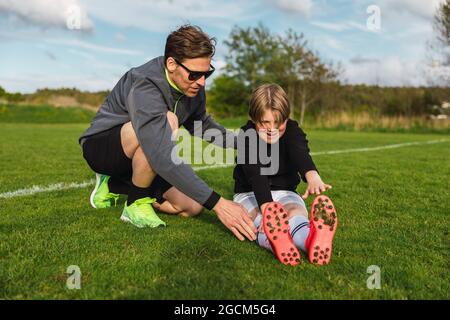 Teen Junge in Sportbekleidung Stretching Beine vor dem Fußballtraining mit Hilfe von männlichen Trainer im grünen Feld Stockfoto