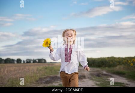 Lächelnder ukrainischer Junge 4-5 Jahre alt in einem gestickten Hemd mit einer Sonnenblumenblume in der Hand läuft vorwärts. Patriotische Erziehung. Symbol der Ukraine, Stockfoto