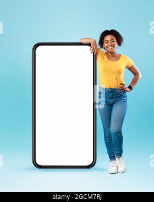 Glückliche schwarze Frau, die sich auf ein großes Smartphone mit einem weißen Bildschirm lehnte, die Kamera auf blauem Hintergrund anlächelte, ein Nachbau Stockfoto