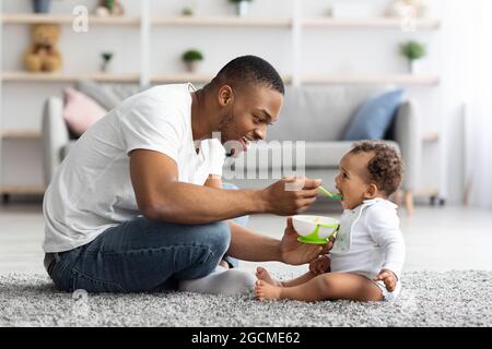Fürsorge des Vaters. Liebevoller Schwarzer Vater Füttert Seinen Niedlichen Baby-Sohn Zu Hause Stockfoto