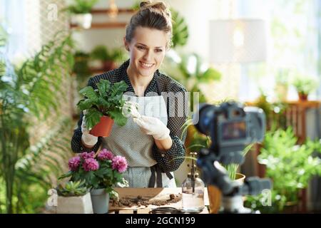 Entspannende Hausgartenarbeit. Glückliche moderne Frau in weißen Gummihandschuhen mit Topfpflanze Aufzeichnungen Gartenarbeit Video-Blog im modernen Haus in sonnigen Tag. Stockfoto