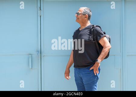 Porträt eines reifen Mannes mit Sonnenbrille und Rucksack, der in den Städtereisen unterwegs ist. Älterer Mann vor einer blauen Tür. Stockfoto