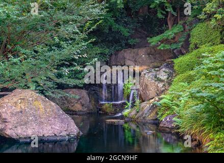 tokio, japan - juli 25 2021: Nahaufnahme Langzeitbelichtung eines kleinen Wasserfalls umgeben von Felsen und Pflanzen im Teich des Hotels New Otani, das für mich berühmt ist Stockfoto
