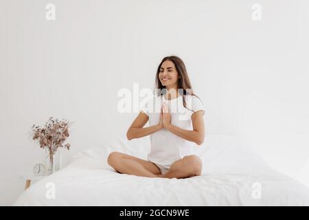 Junge lächelnde Frau, die zu Hause Yoga auf dem Bett praktiziert. Stockfoto