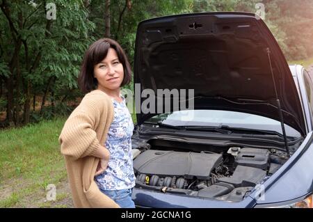 Eine junge Frau, die in der Nähe eines kaputten Autos mit angehobener Haube steht, hat Probleme mit ihrem Fahrzeug. Traurige verärgerte Frau. Stockfoto