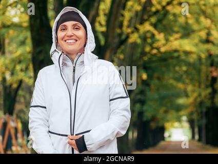 Mit freundlichen Grüßen lächelnd Fit athletische Frau trägt eine moderne wasserdichte Laufjacke mit bunt grün gelben Herbst Park Hintergrund. Aktive Menschen konce Stockfoto