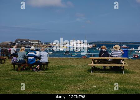 Menschen, die auf Picknicktischen in Hugh Town sitzen, mit der Scillonischen Fähre in der Ferne, St. Mary's, Isles of Scilly, Cornwall, England, Großbritannien, Juli 2021 Stockfoto