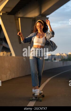 Junge unbeschwerte Frau genießt es, bei Sonnenuntergang Skateboard zu fahren. Glückliches Mädchen in trendigen Street-Style-Kleidung Stockfoto