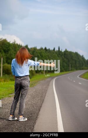 Das Mädchen hält das Auto mit der Hand auf der Autobahn an. Stilvolle Frau auf der Straße hält das Auto auf eine Reise gehen. Eine Straße mitten im Wald. Stockfoto