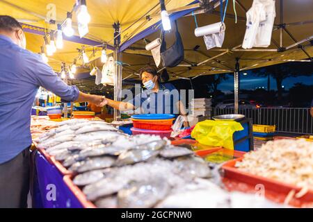 Street Food Nachtmarkt in Putrajaya, in der Nähe von Kuala Lumpur. Verkäufer mit Gesichtsmaske in einem Seafood Street Store. Der Kunde zahlt für den Fisch, den er kauft Stockfoto