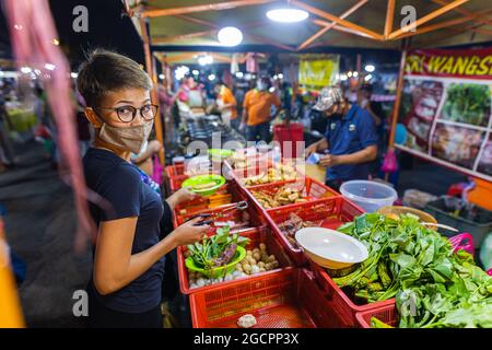 Street Food Nachtmarkt Putrajaya, in der Nähe von Kuala Lumpur. Junge asiatische Mädchen kauft Gemüse in einem Nachtmarkt. Malaysische Frauen mit Gesichtsmaske in einer Straße Stockfoto