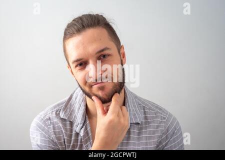 Der junge kaukasische Mann starrt fröhlich an seinem Kinn in grauem Hemd gegen weißen Hintergrund kratzen Stockfoto