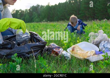 Weibliche Hände in gelben Gummihandschuhen sammeln Plastikmüll in einem schwarzen Beutel im Freien. Im Hintergrund hilft ein Junge. Selektiver Fokus. Sommer sonnig Stockfoto