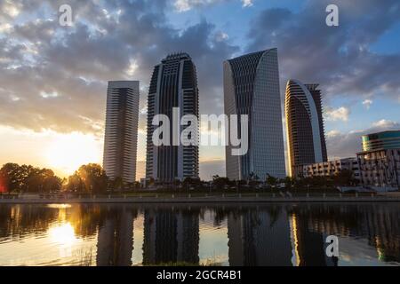 Wolkenkratzer bei Sonnenuntergang, vor einem See mit Spiegelungen der Gebäude. Wunderschöner farbenprächtiger Sonnenuntergang hinter Hochhäusern. Geschäftsviertel illumina Stockfoto