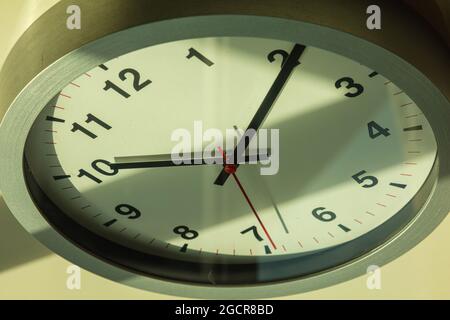Eine moderne weiße Wanduhr im Sonnenlicht mit Schatten auf dem Zifferblatt. Nahaufnahme einer Wanduhr mit einem roten Sekundenzeiger. Die Zeiger auf 10 nach Stockfoto