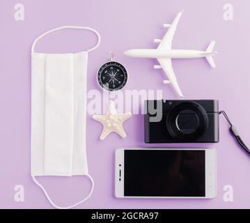 Welttourismustag, Ansicht von oben von minimalem Modellflugzeug, Flugzeug, Seesterne, Kompass, Smartphone leerer Bildschirm und Gesichtsmaske isoliert auf violettem Hintergrund, Stockfoto