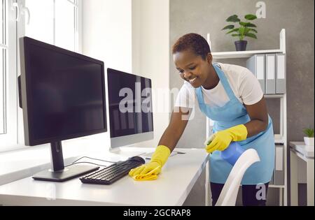 Fröhlicher lächelnder Reiniger wischt den Staub in einem hellen Büro vorsichtig vom Tisch. Stockfoto