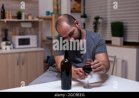 Kaukasischer Mann hält ein Glas Wein in der Küche zu Hause. Depressive Person, die Alkohol trinkt, Alkohol trinkt, alkoholisches Getränk allein, während sie sich mit einer Flasche Alkohol berauscht fühlt Stockfoto