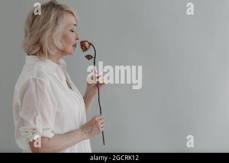 Traurige ältere Frau im weißen Hemd hält eine trockene, alte, tote Rosenblume. Alter und Alterungskonzept. Stockfoto