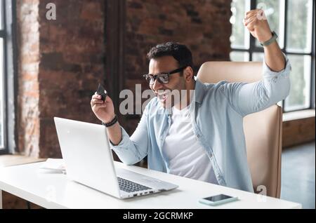 Ein junger, überfröhlicher Geschäftsmann mit gemischtem Rennen, der mit offenem Mund vor einem Laptop am Schreibtisch sitzt, eine Kreditkarte hält und sich über eine erfolgreiche Transaktion oder den erheblichen Preisnachlass freut Stockfoto