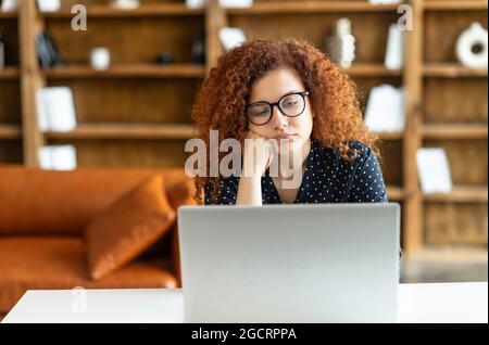 Junge Rothaarige lockige Frau mit Laptop sitzt im Büro und fühlt sich gelangweilt, Müdigkeit, Mangel an Motivation und Energie, weibliche Mitarbeiterin müde von nicht interessanten und monotonen Arbeit Stockfoto