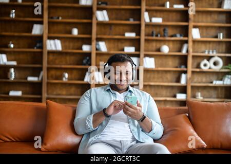 Ein lächelnder Inder mit Kopfhörern sitzt mit einem Smartphone auf dem Sofa, spielt Videospiele, hört Musik, schaut sich Videos oder Filme an. Fröhlicher Ostmann mit Ohrhörern in legerer Kleidung hält das Telefon Stockfoto