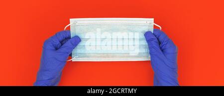 Die Hände des Arztes in Handschuhen halten eine medizinische Gesichtsmaske, ein Banner mit einem Gesundheits- und Hygienekonzept auf rotem Hintergrund Stockfoto