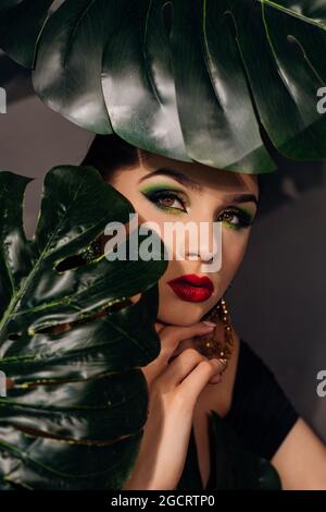 Nahaufnahme des Gesichts einer schönen jungen Frau, die sich mit einer grünen Monstera-Palme bedeckt. Portrait der Schönheit Frau mit Make-up rote Lippen klare Haut mit großen grünen Stockfoto