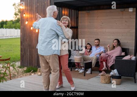 Glückliches Seniorenpaar, das vor der Familie auf der Terrasse des Landhauses tanzt Stockfoto