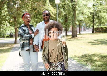 Netter glücklicher Schuljunge, der am sonnigen Tag vor seinen Eltern auf der Straße im Park unterwegs ist Stockfoto