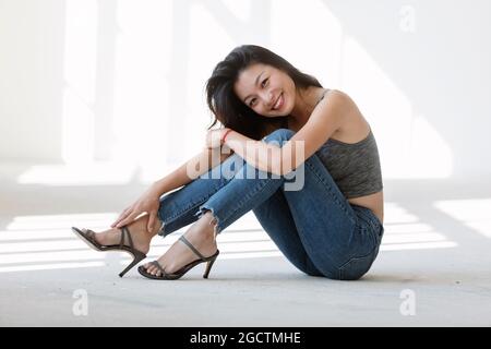Portrait schöne asiatische Mädchen auf dem Boden sitzen. Sie trägt blaue Jeans, ein sportliches Top und Stiletto Heels. Dahinter kommt das Sonnenlicht.