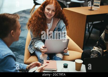 Mädchen mit Laptop im Gespräch mit Freund. Junge Frau mit Laptop sitzt mit Freunden in der Universitätsbibliothek. Stockfoto