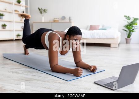 Plus size Afro Dame, die zu Hause mit Online Personal Trainer trainiert, mit Laptop, stehend in Ellenbogenplankenpose Stockfoto