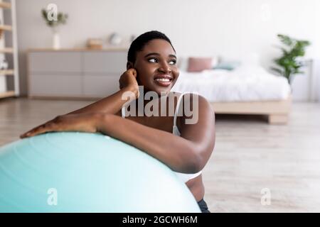 Fröhliche übergewichtige schwarze Frau, die sich auf den Fitnessball stützt und in Innenräumen Sport macht Stockfoto