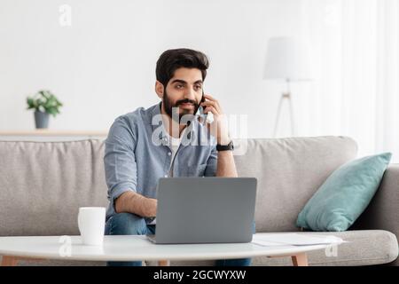 Immer in Kontakt. Arabischer Freiberufler, der mit einem Laptop auf dem Handy spricht, der aus der Ferne arbeitet, auf dem Sofa sitzt und freien Platz hat Stockfoto