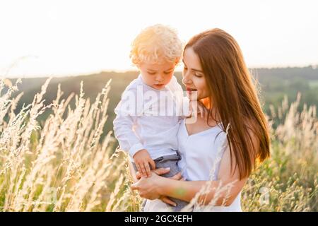 Eine wunderschöne Frau mit einem kleinen, lockigen Jungen in den Armen steht bei Sonnenuntergang mitten auf einem Feld Stockfoto