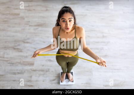 Oben Blick auf die schockierte indische Dame auf der Waage, Überprüfung der Taillenmaße, überrascht mit Gewichtsverlust Ergebnisse Stockfoto