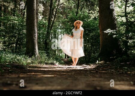 Ein Mädchen in einem weißen Kleid und Hut geht durch einen Sommerpark oder Wald. Märchenwald. Foto in voller Länge von glücklicher Frau Stockfoto