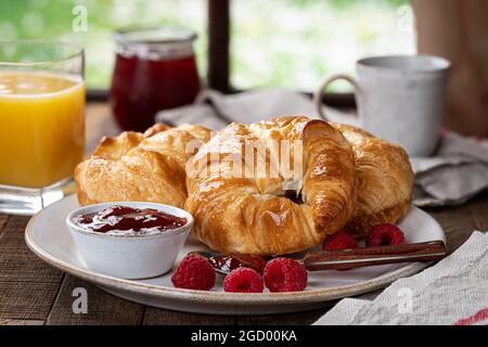 Croissants mit Himbeerkonfitüre und frischen Himbeeren auf einem Teller am Fenster Stockfoto