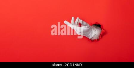 Santa Claus Hand trägt weißen Handschuh durch ein Loch in rotem Papier Hintergrund Stockfoto