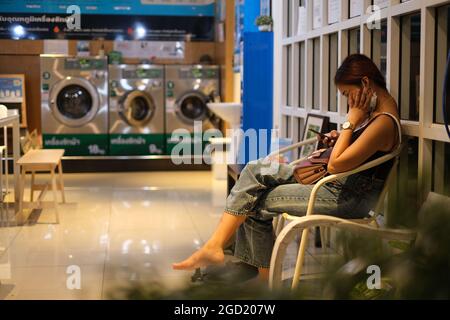 Eine junge Thailänderin liest Smartphone, während sie in einer Wäscherei darauf wartet, dass ihre Kleidung gewaschen wird Stockfoto
