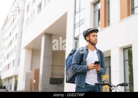 Mittellange Aufnahme eines seriös schönen männlichen Kuriers mit Thermorucksack, der mit dem Fahrrad auf der Stadtstraße steht und die Navigations-App am Telefon nutzt. Stockfoto