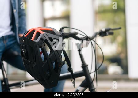 Nahaufnahme des Fahrradhelms, der am Lenker des Fahrrads hängt, das am Sommertag auf der Stadtstraße steht, verschwommener Hintergrund des städtischen Gebäudes. Stockfoto