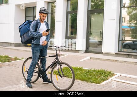 Mittellange Aufnahme eines gutaussehenden männlichen Kuriers mit Thermorucksack, der mit dem Fahrrad auf der Stadtstraße steht und die Navigations-App am Telefon nutzt. Stockfoto